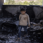 Boy stands amid charred debris in a burned, ruined room, with blackened walls and scattered rubble around him.