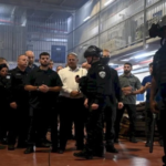 Group of uniformed security officers in a workshop-like setting, listening to a demonstration; two officers in the foreground wear helmets and protective gear.