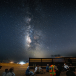 Milky Way stretches across a star-filled night sky above a desert gathering, with people seated near a wooden barrier and a person in a reflective vest leading the event.