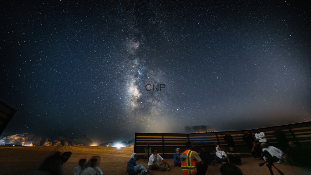 image - Center News Network Milky Way stretches across a star-filled night sky above a desert gathering, with people seated near a wooden barrier and a person in a reflective vest leading the event.