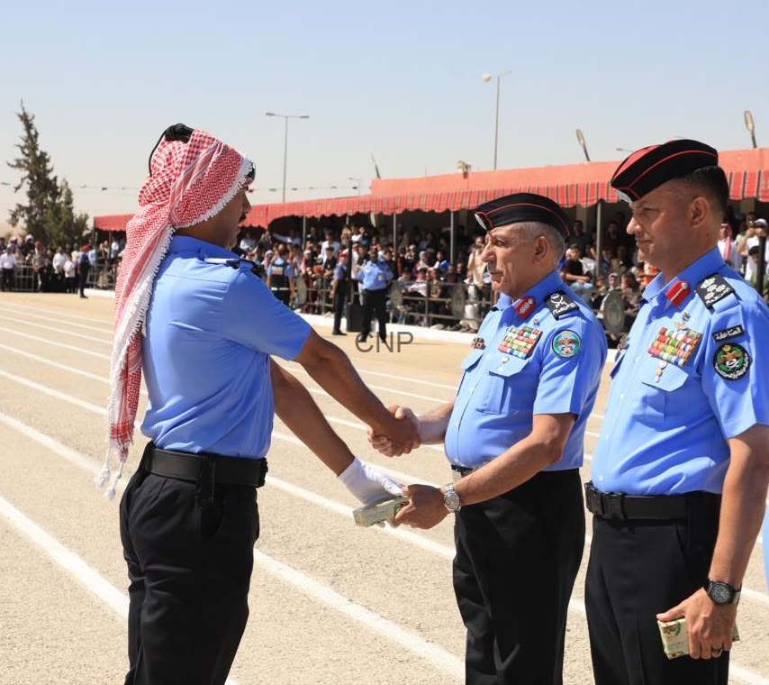 Man in a blue uniform with a red-and-white keffiyeh shakes hands with a senior officer on a running track during a ceremony, with spectators under red canopies in the background.
