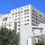 White stone Ministry of Health building with an arched entrance, palm trees, and a bright blue sky; appears to be a government health complex.