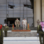 Military officers stand on a wooden platform at the top of stone stairs in front of a glass-fronted building during a formal ceremony, with guards in the foreground.