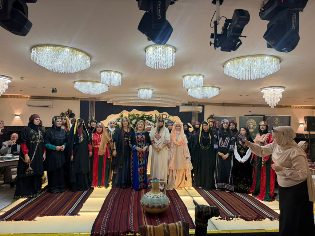 Group of women in colorful traditional dresses posing together on a stage at a cultural event, chandeliers overhead.