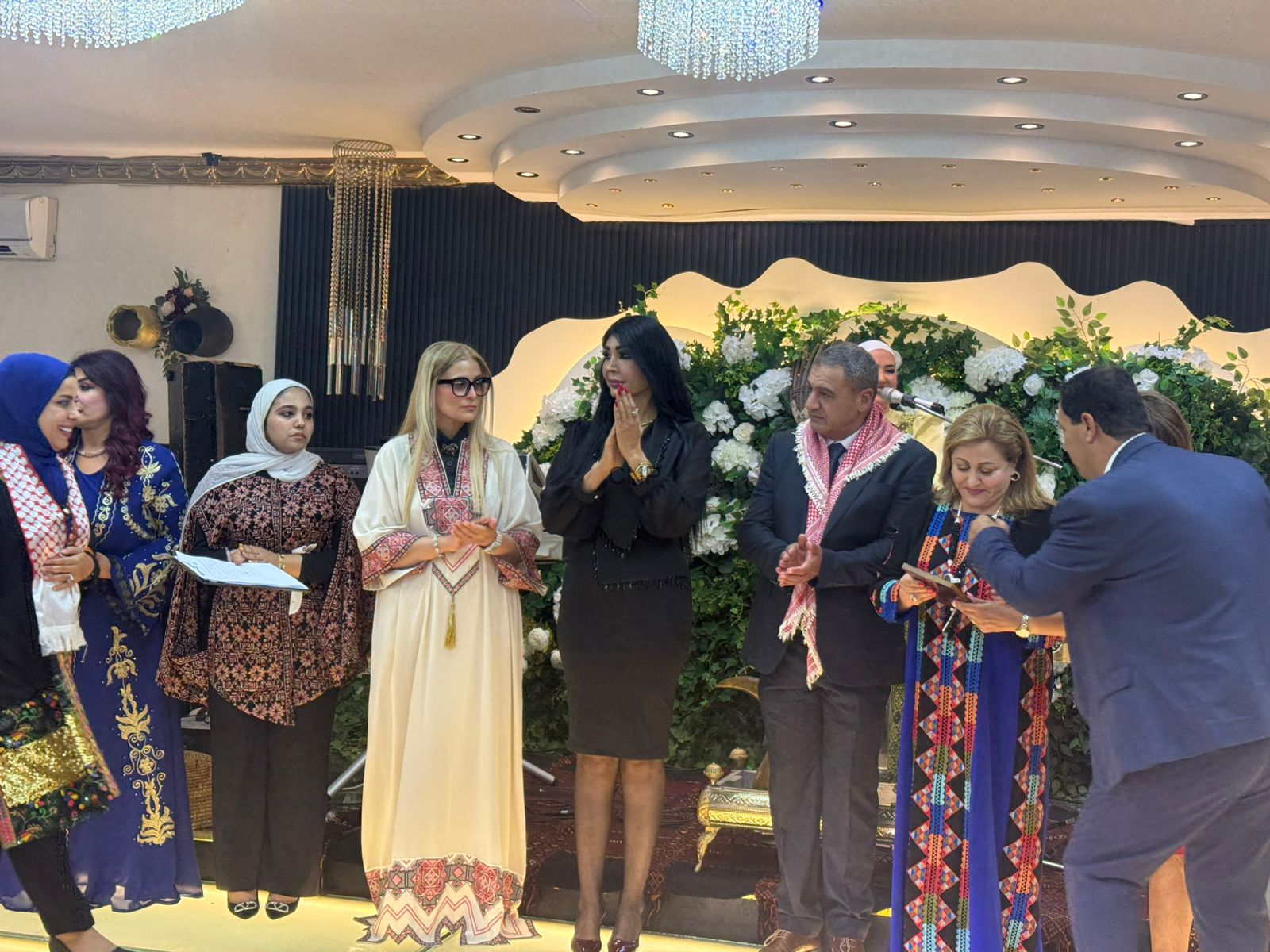 image - Center News Network Group of people in traditional dresses on a decorated stage during a ceremony, with white flowers and greenery in the background.