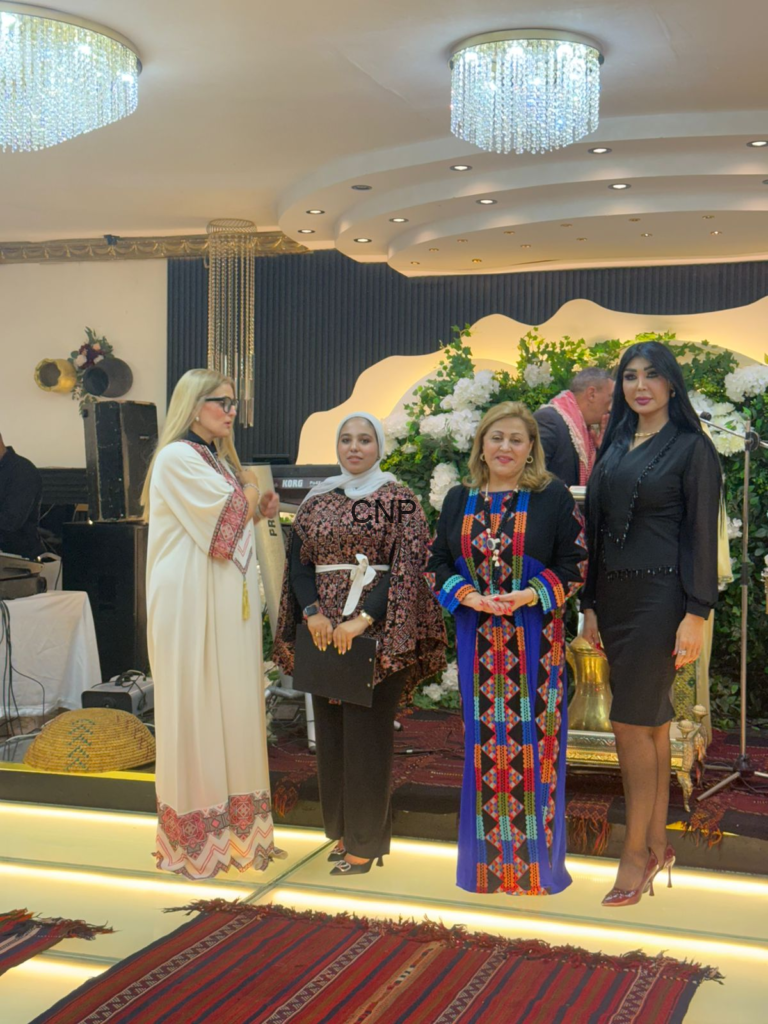 Four women in colorful traditional outfits posing on a stage with a floral backdrop at an indoor event.
