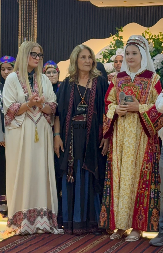 Group of women in traditional garments on stage, one holding a microphone, others in colorful robes with patterns and head coverings.