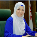 Woman in a white hijab and blue outfit seated at a conference table, looking at the camera.
