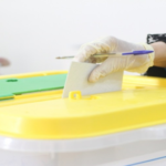 Person wearing gloves inserts a ballot into a yellow ballot box during voting.