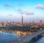 Aerial view of a coastal city with a long bridge across a bay and a dense skyline under a blue sky with clouds; watermark 'CNP'.