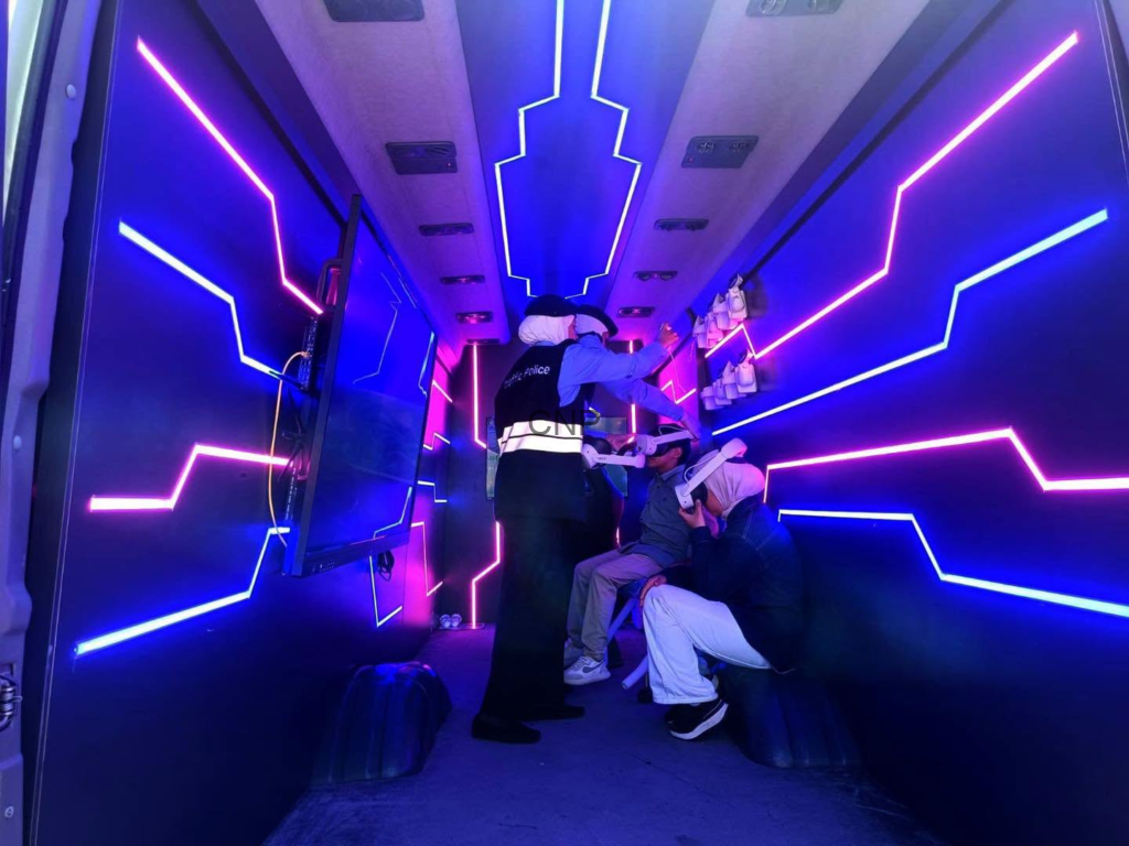 Inside a neon-lit vehicle, a uniformed officer in a reflective vest talks to two seated passengers while others stand nearby.