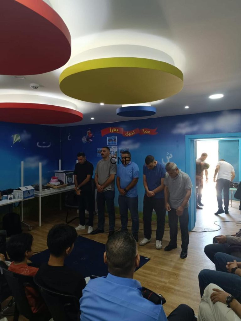 Five men stand with heads bowed in a line inside a bright blue room, audience seated and observing, colorful circular ceiling lights overhead.