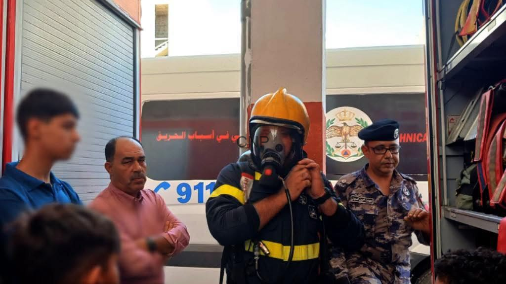 image - Center News Network Firefighter in a yellow helmet and gas mask speaks into a microphone beside an ambulance, while a uniformed officer and bystanders look on.