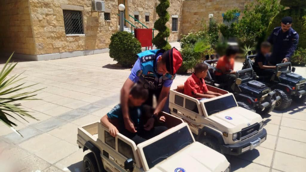 Police officer in uniform helps children ride in small plastic toy SUVs in a sunny courtyard with beige stone buildings and greenery.