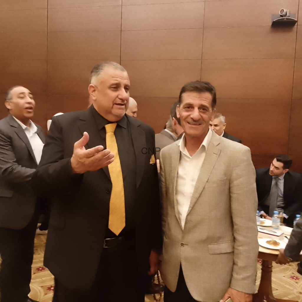 image - Center News Network Two men in suits pose for a photo at a formal event; the man on the left gestures with his hand as the other smiles at the camera in a banquet hall setting.