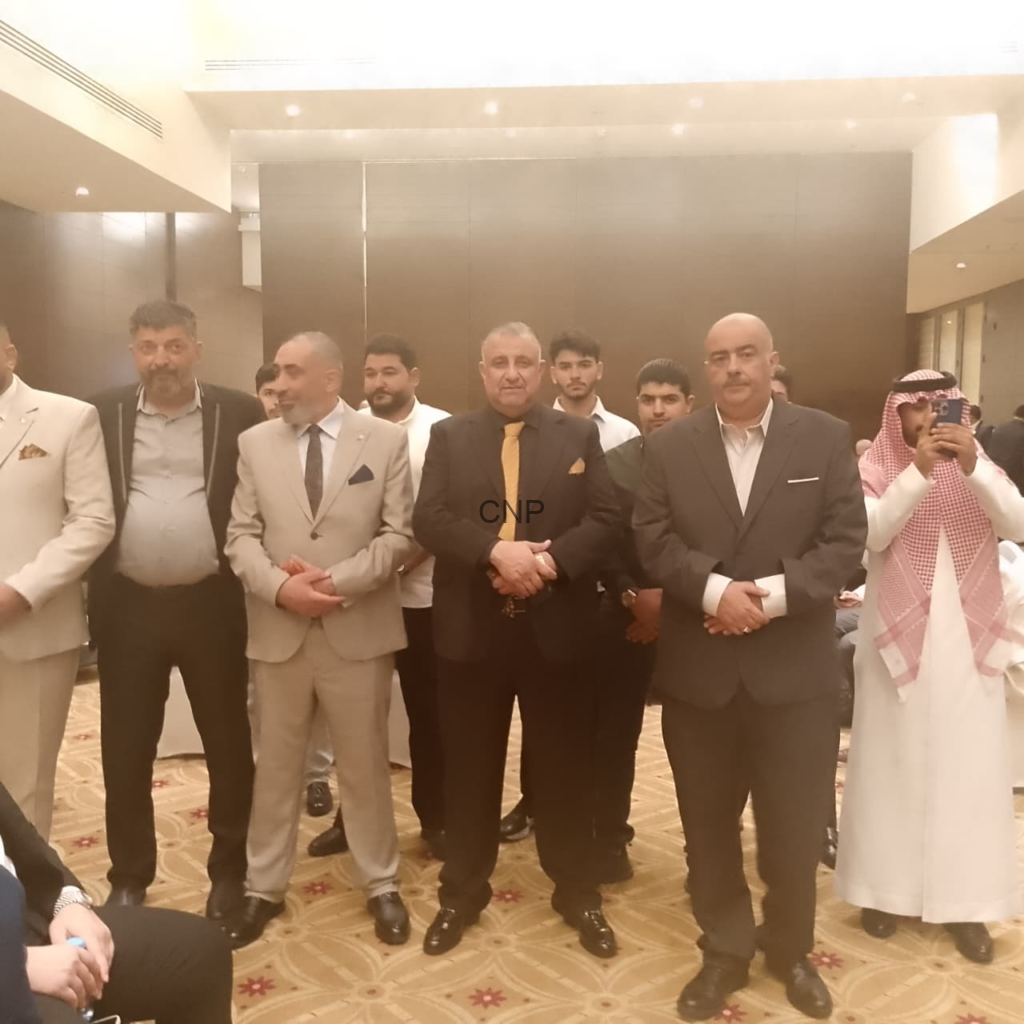 Group of men in business suits posing for a photo in a banquet hall during an event, with one man in traditional attire on the far right taking a picture.