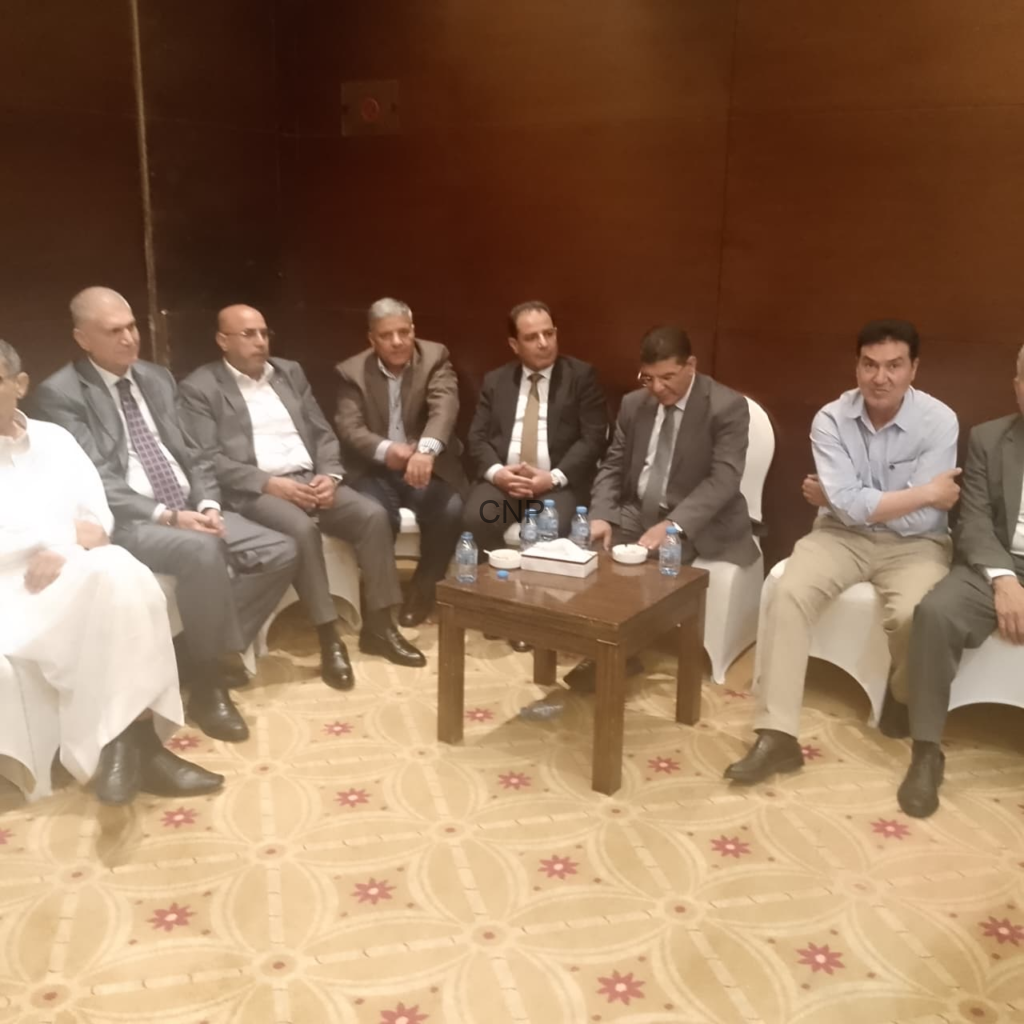 Group of men in suits seated in a row around a small table with water bottles, in a formal meeting setting.