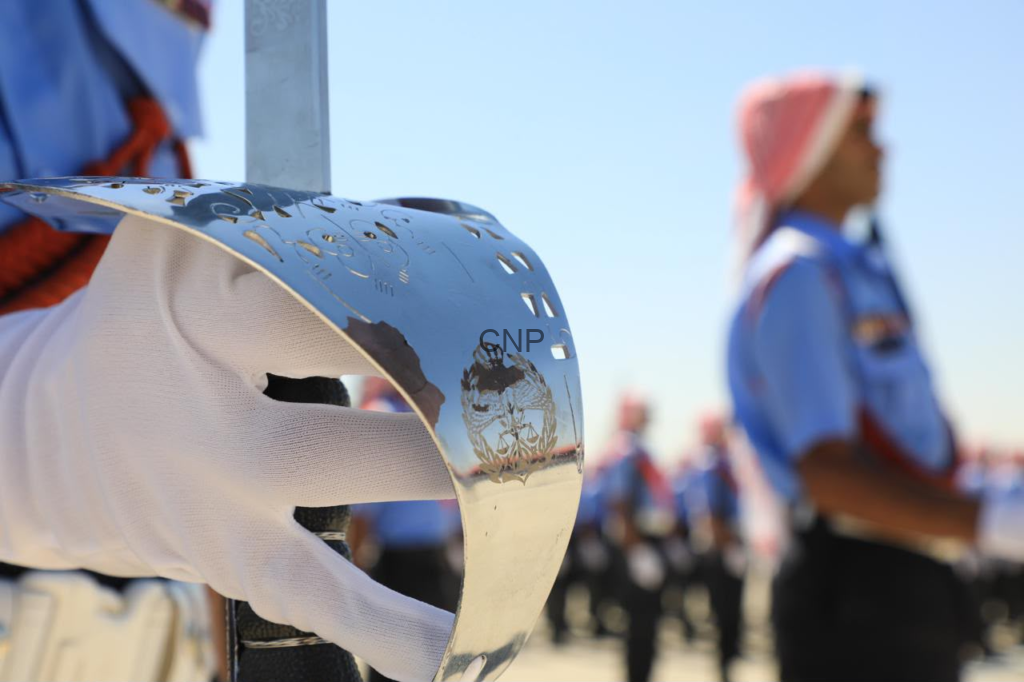 Close-up of a shiny metal ceremonial helmet held by a white-gloved hand during a parade, with uniformed marchers blurred in the background.
