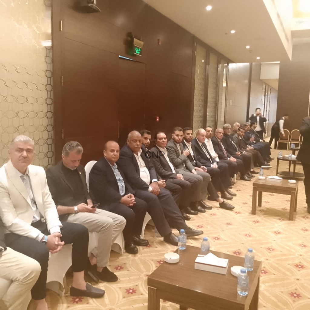 Row of men in suits seated along a wall in a hotel conference room, with tables and water bottles in the foreground.