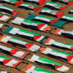 Dozens of white stretchers draped with Palestinian flags arranged in neat rows on sandy ground, likely for a funeral or memorial ceremony.