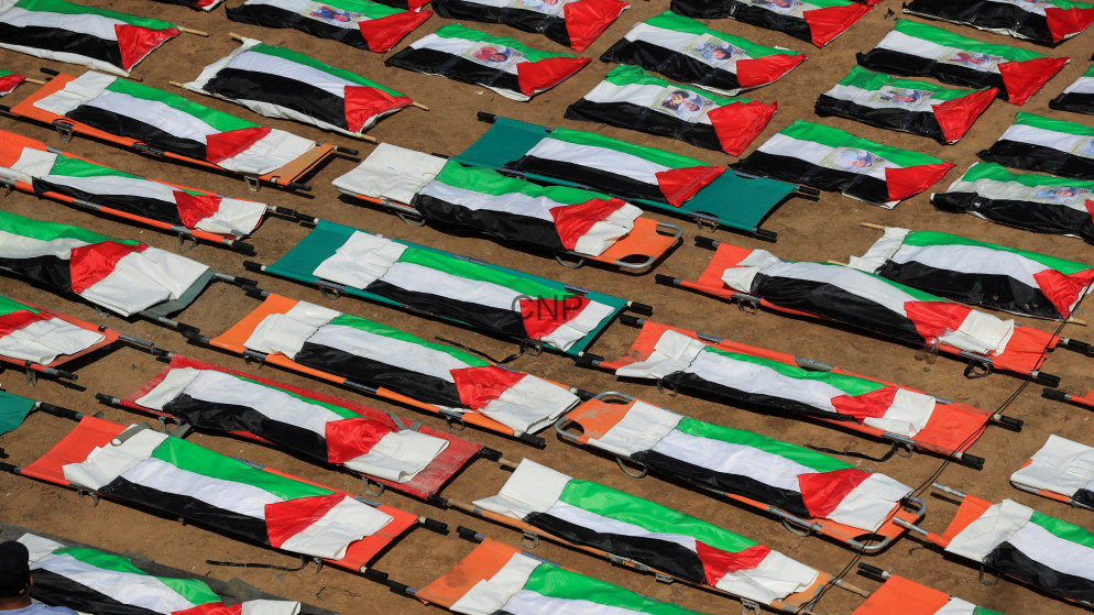 Dozens of white stretchers draped with Palestinian flags arranged in neat rows on sandy ground, likely for a funeral or memorial ceremony.