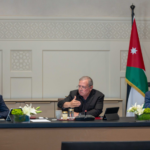 Three men sit at a conference table with Jordanian flags on either side; center man speaks while the others listen.