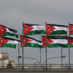 Several Jordanian flags fluttering on poles atop a building roof against a cloudy sky.