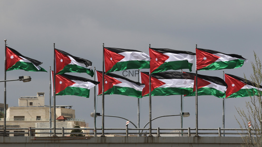 Several Jordanian flags fluttering on poles atop a building roof against a cloudy sky.