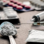 Spoon with white powder and a syringe on a table, with pill blister packs in the background, implying drug use.