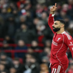 Liverpool forward Mohamed Salah raises his right index finger in celebration on the pitch with a blurred crowd behind him.
