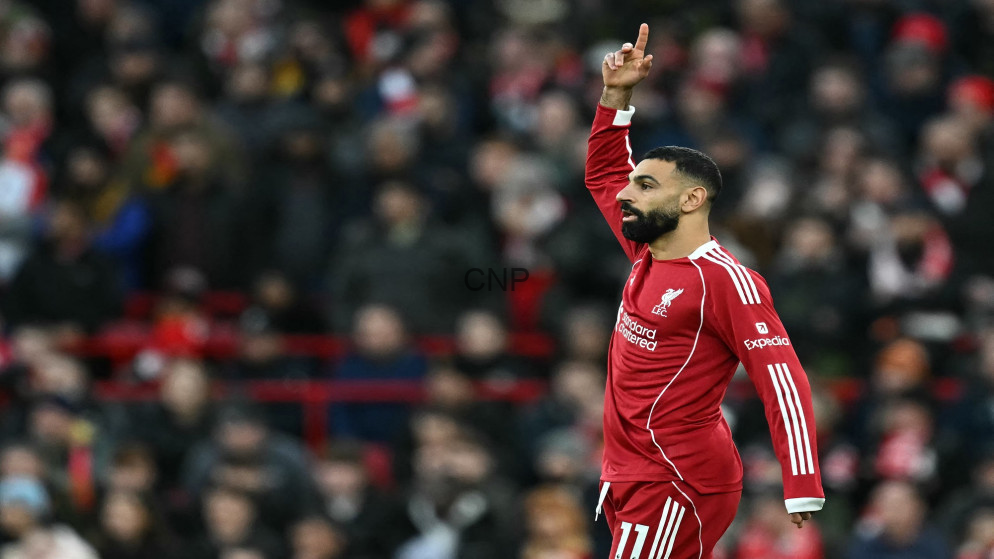 Liverpool forward Mohamed Salah raises his right index finger in celebration on the pitch with a blurred crowd behind him.