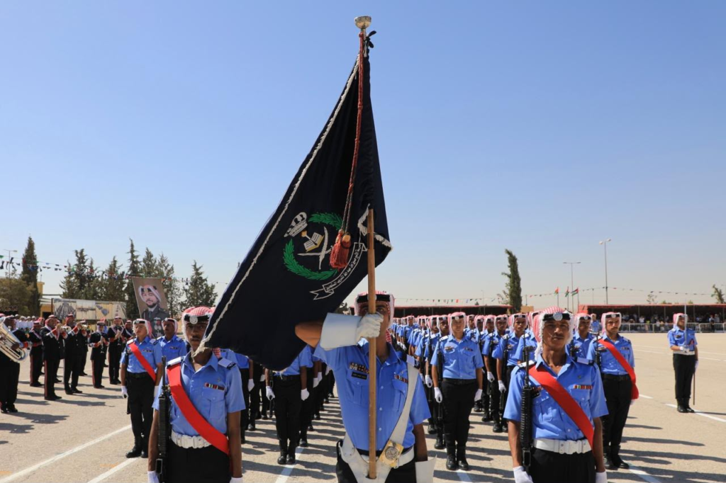 image - Center News Network Color guard leading a parade: flag bearer holding a large black banner with emblem, surrounded by rows of uniformed troops in blue shirts and red sashes on a sunny day.