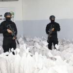 Two armed guards in black tactical gear stand among white bags in a warehouse, each holding a rifle.