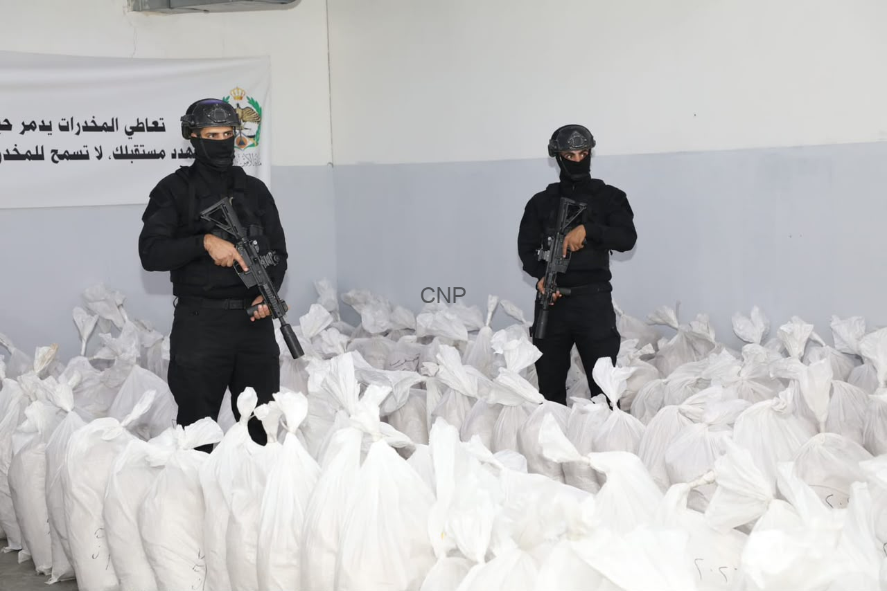 image - Center News Network Two armed guards in black tactical gear stand among white bags in a warehouse, each holding a rifle.