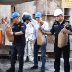 Group of workers in hard hats standing near a furnace, handing out brown paper bags to each other.