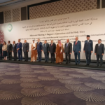 Group of officials in formal attire standing on a stage for a ministerial meeting photo, with a white banner and emblem on the wall behind them.