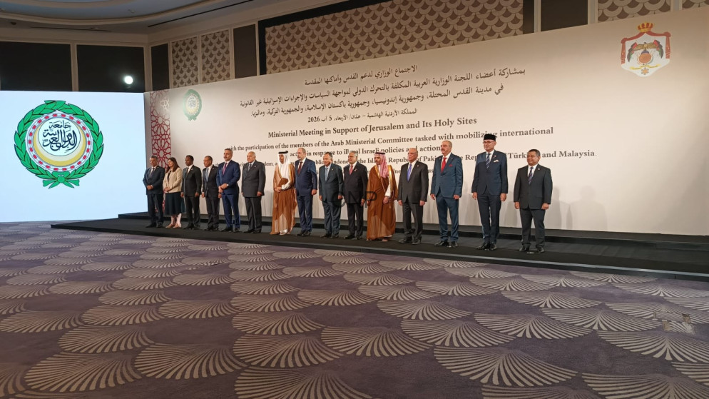 Group of officials in formal attire standing on a stage for a ministerial meeting photo, with a white banner and emblem on the wall behind them.