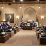 Diplomatic meeting in a beige, opulent room with two leaders seated center, flags behind them, and delegates on both sides.