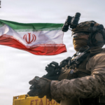 Armed soldier in desert camouflage with night-vision goggles, standing beside an Iranian flag.