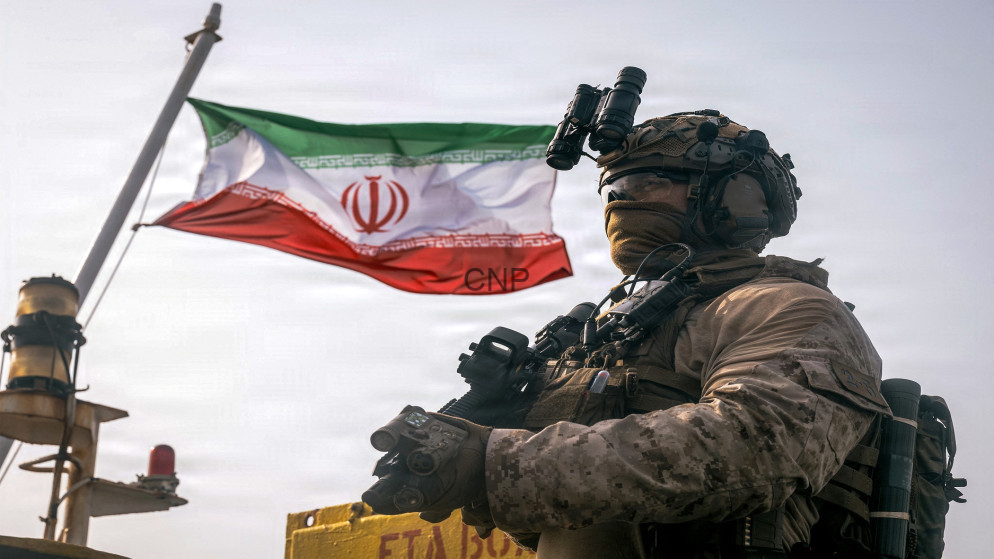 Armed soldier in desert camouflage with night-vision goggles, standing beside an Iranian flag.