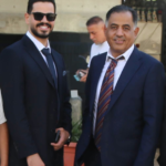 Two men in business suits posing for a photo outdoors; the man on the left wears sunglasses and a dark suit.