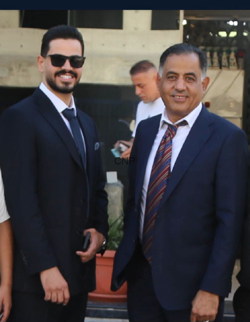 Two men in business suits posing for a photo outdoors; the man on the left wears sunglasses and a dark suit.