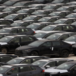Row after row of dark sedans parked in a car lot, many with protective white covers on hoods and windows