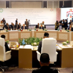 Large formal international meeting with delegates seated around a long U-shaped table, national flags and media onlookers nearby, stage in background.