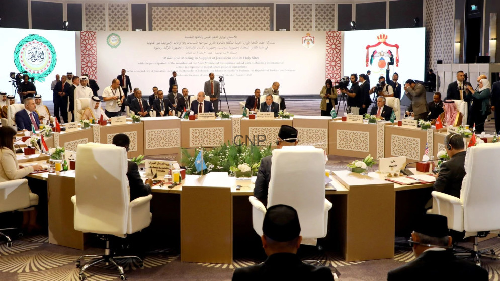 Large formal international meeting with delegates seated around a long U-shaped table, national flags and media onlookers nearby, stage in background.