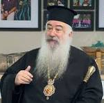 Orthodox priest in black vestments and hat with a gold cross, giving a thumbs-up indoors, framed pictures on the wall behind.