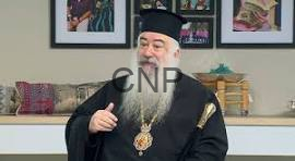 Orthodox priest in black vestments and hat with a gold cross, giving a thumbs-up indoors, framed pictures on the wall behind.