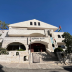 Stone public building with arched entrance, two trees, and a Jordanian flag on a sunny street.