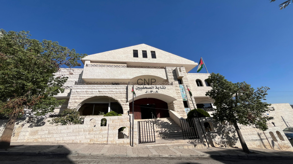Stone public building with arched entrance, two trees, and a Jordanian flag on a sunny street.