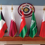 Seven national flags on a wooden podium with gold finials, arranged in a row in front of a wall with a circular seal.